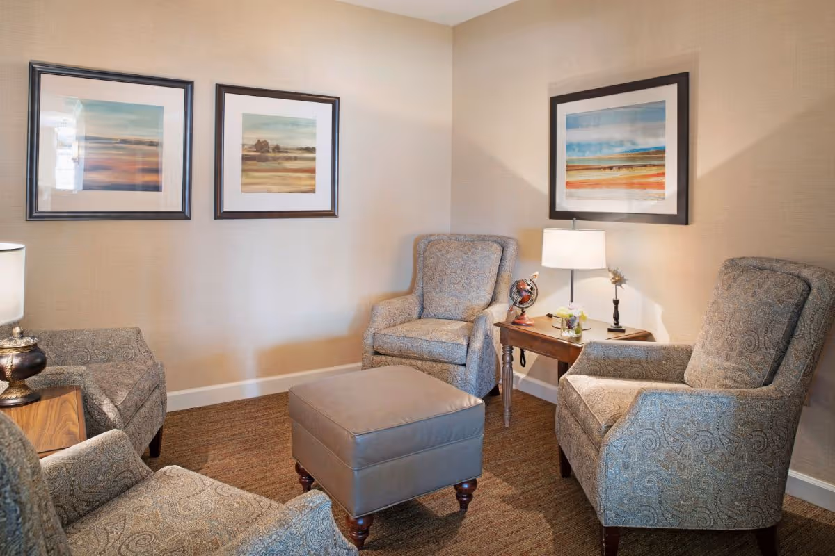 A cozy sitting area with four patterned armchairs arranged around a square ottoman. There are two wooden side tables with lamps and decorative items, and three framed landscape paintings on the beige walls.