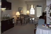 Interior view of a living area in Avon Oaks Caring Community featuring a small kitchenette with a sink and microwave on the left, a striped sofa with two table lamps behind it, a coffee table, an armchair, and a round table with a tablecloth and decorative items in the foreground. A window and a door with blinds are visible in the background.