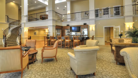 Spacious and well-lit senior living facility common area with comfortable armchairs and sofas arranged around small tables, a central bar with high chairs, a staircase leading to an upper floor balcony, and warm lighting fixtures on the walls.