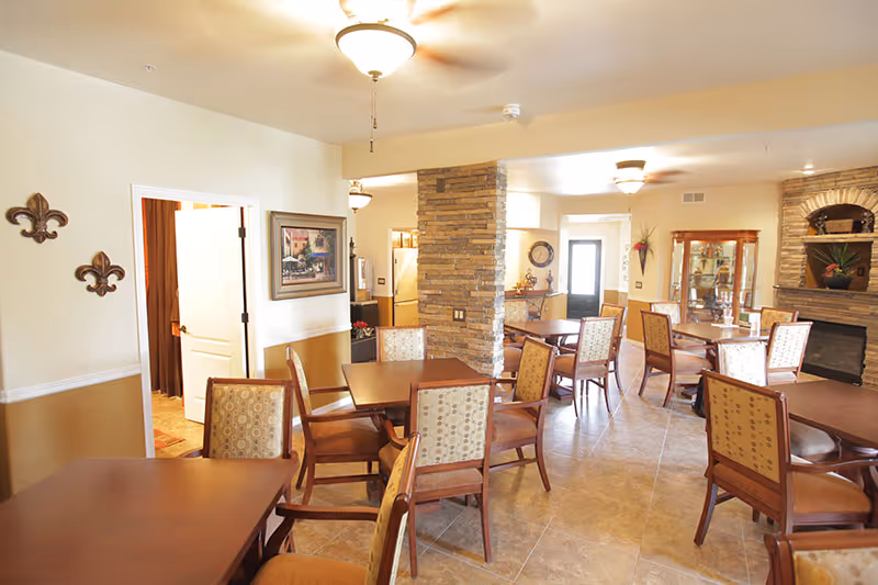 A bright and spacious dining area in a senior living facility with multiple wooden tables and cushioned chairs arranged neatly. The room features a stone pillar in the center, a fireplace with stone surround, a glass display cabinet, and wall decorations including framed artwork and fleur-de-lis ornaments. The floor is tiled, and ceiling fans with lights are mounted on the ceiling.