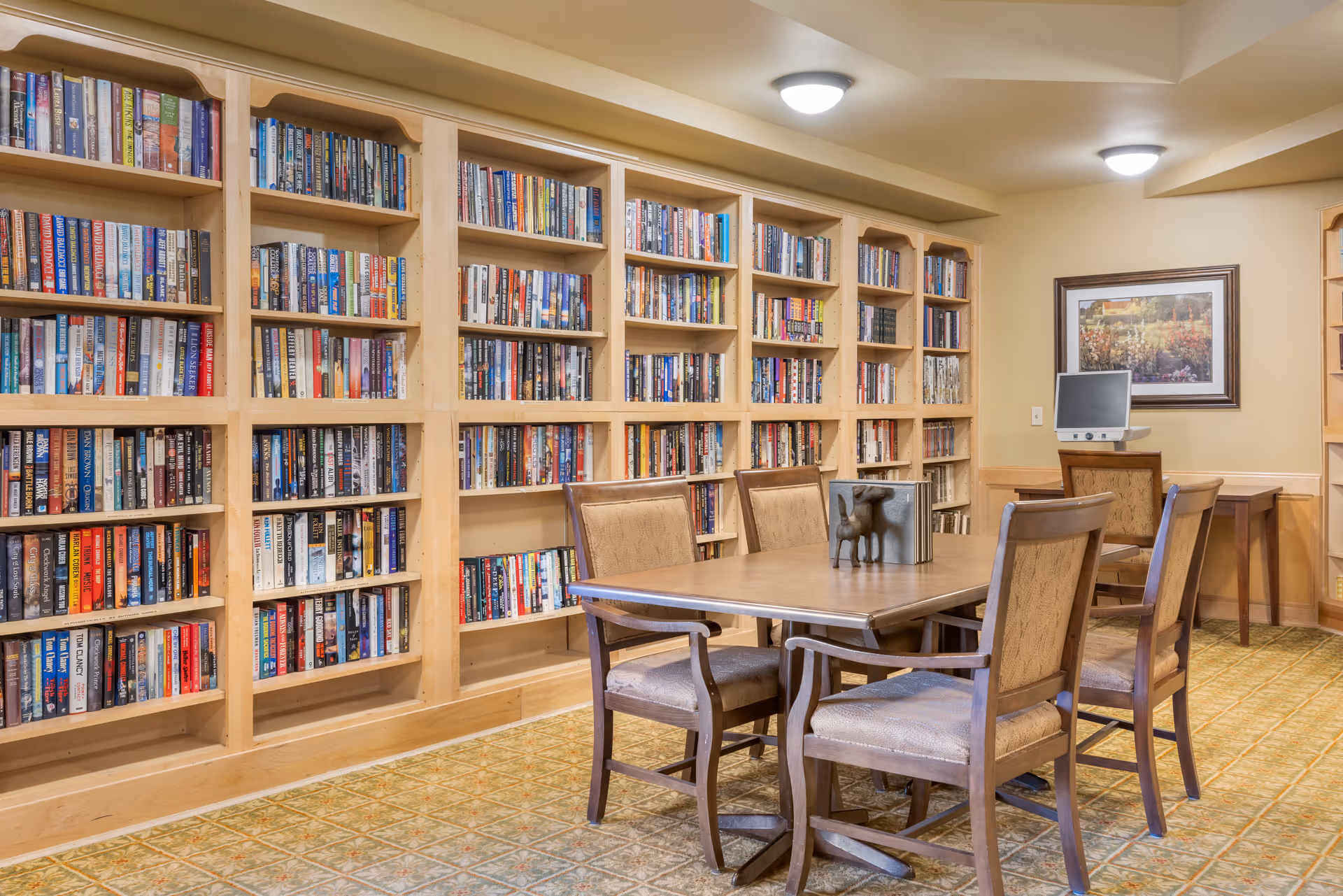 A cozy library room with wooden bookshelves filled with books covering the entire wall. In the center, there is a wooden table surrounded by four cushioned chairs. On the table, there is a decorative item and a few books. In the background, there is a small desk with a computer and a framed picture hanging on the wall. The room has warm lighting and a patterned carpeted floor.