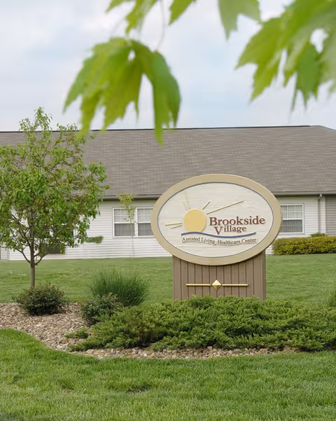 A wooden sign reading "Brookside Village" stands on a landscaped lawn in front of a single-story senior living building with trees and overhanging leaves.