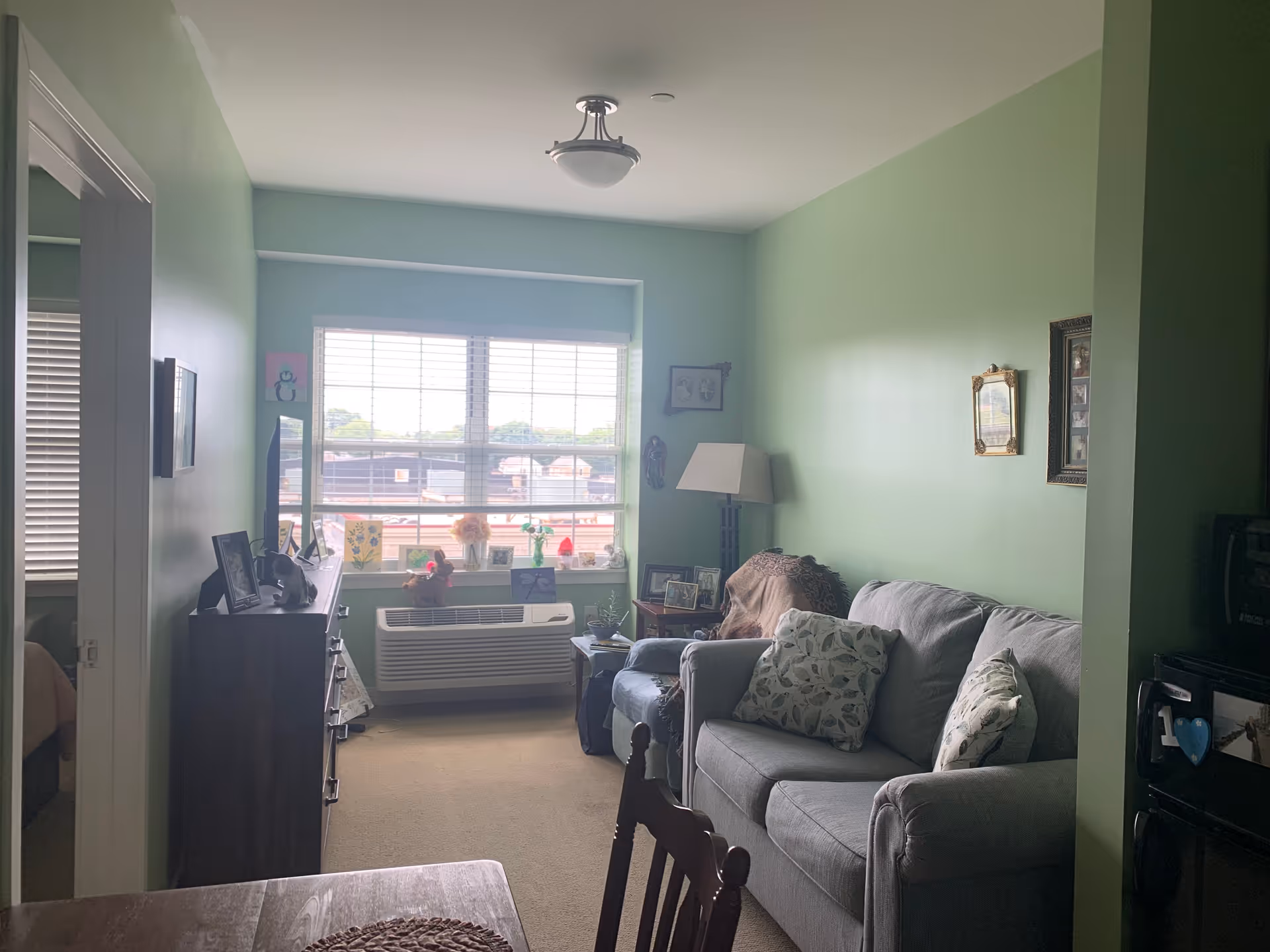 A cozy living room with light green walls, a gray couch with patterned pillows, a blue armchair, a floor lamp, and a window with blinds letting in natural light. There is a wooden dresser with framed photos and decorations, and a small table with a chair in the foreground. The room has a carpeted floor and some wall art.