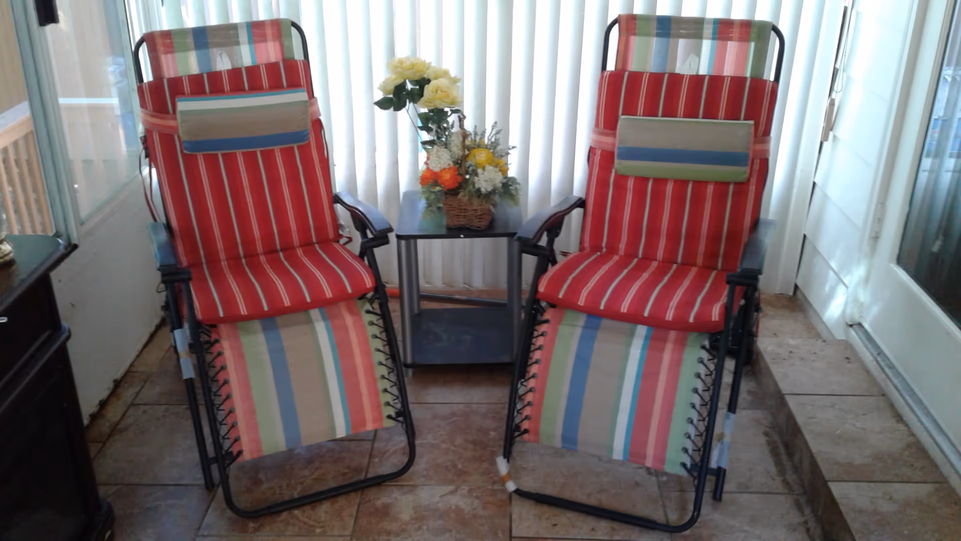 Two reclining chairs with red and multicolored striped cushions are positioned side by side on a tiled floor. Between the chairs is a small black table with a basket of artificial flowers. Vertical blinds cover the window behind the chairs, and a glass door is visible on the right side.