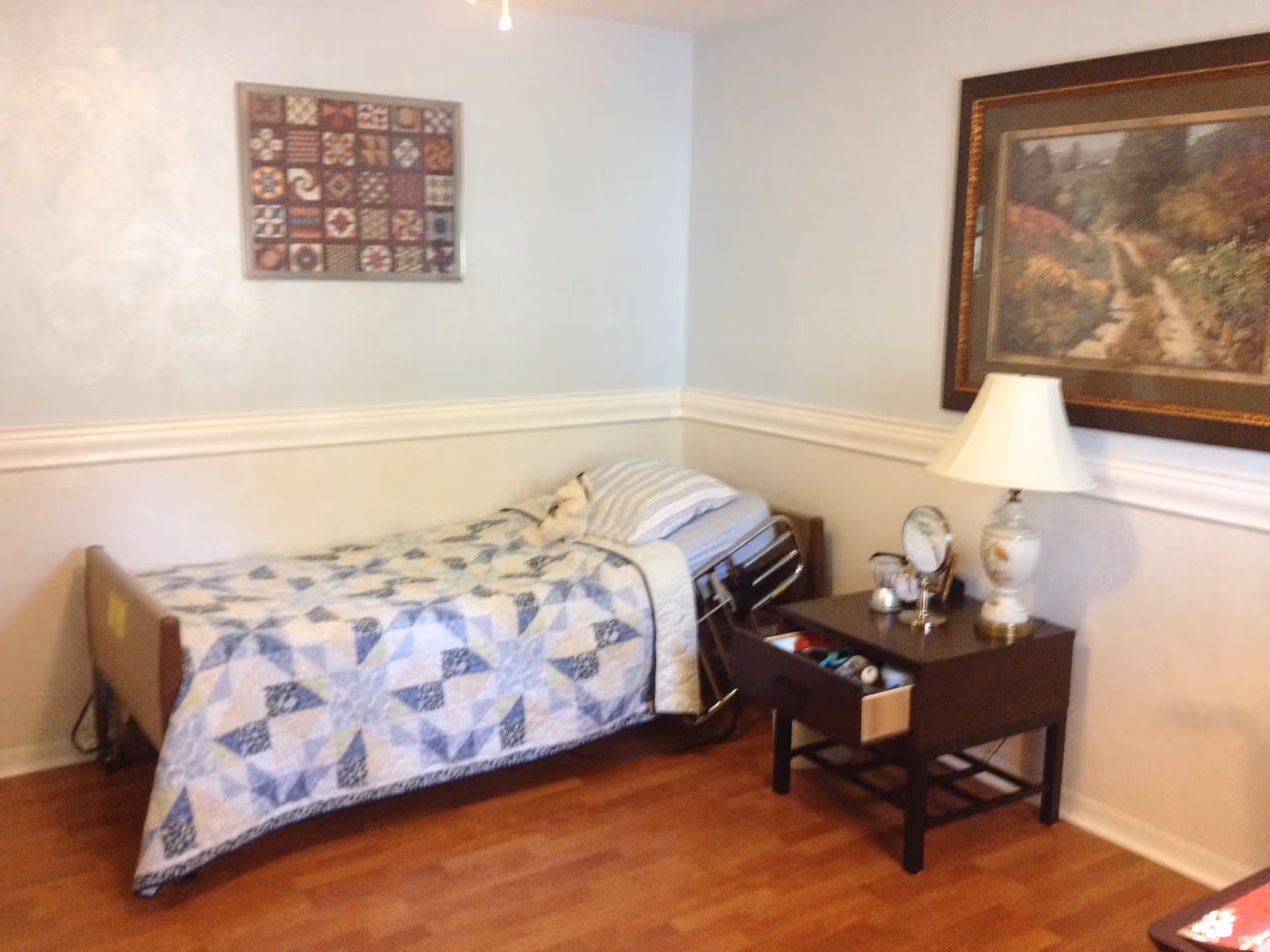 A small bedroom with a single bed covered with a blue and white patterned quilt. Next to the bed is a dark wooden nightstand with an open drawer containing various items, a table lamp, and a clock. The walls are light-colored with a chair rail molding, and two framed pictures hang on the walls. The floor is wooden.