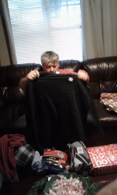 An elderly person sitting on a dark brown leather couch holding up a black sweater. In front of them on a table are various wrapped gifts and clothing items. Behind them is a window with white blinds and light-colored curtains.