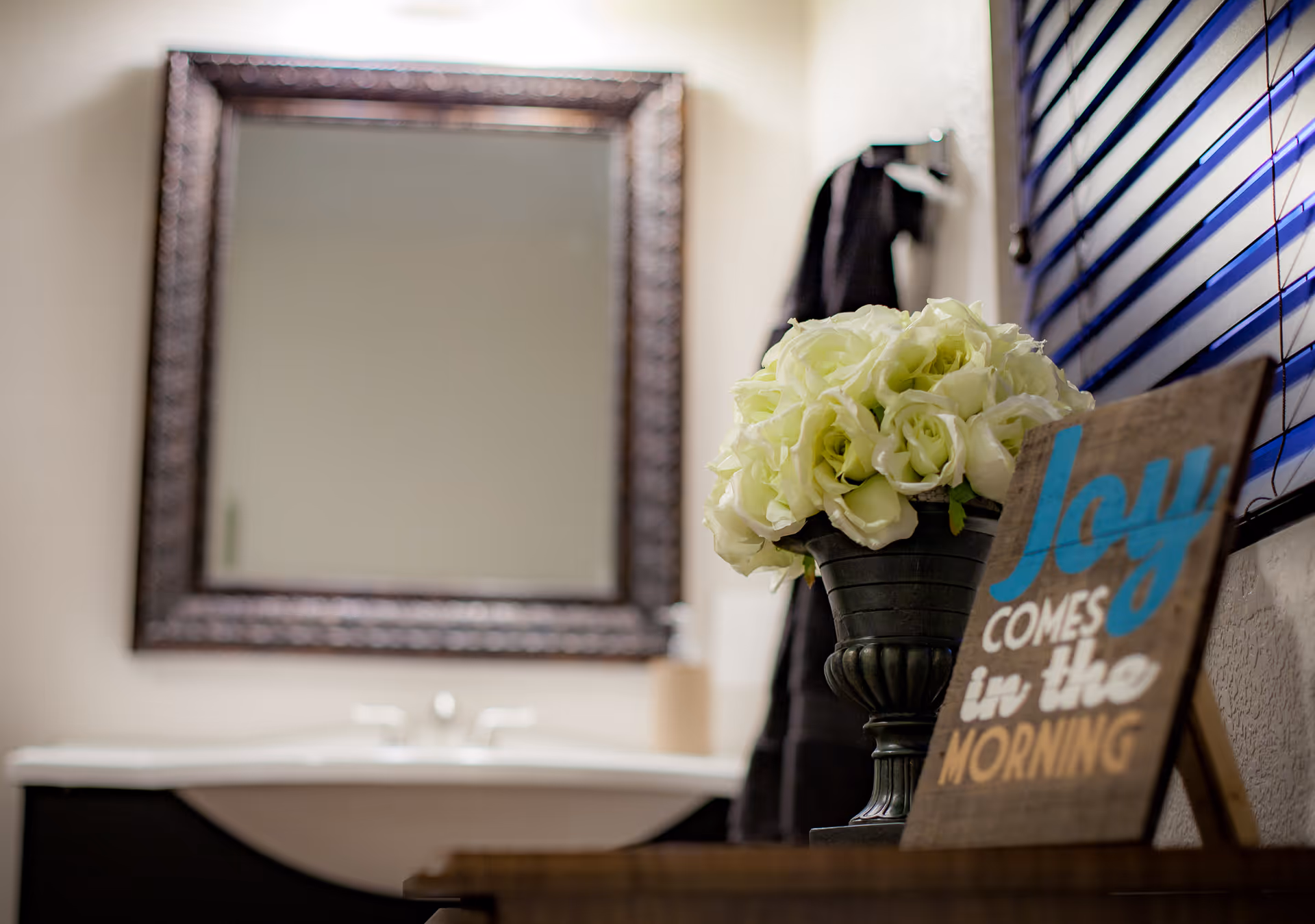 A bathroom interior featuring a sink with a large ornate framed mirror above it. In the foreground, there is a vase with white flowers and a decorative sign that reads 'Joy comes in the morning.' A black towel hangs on a rack next to a window with blue blinds.