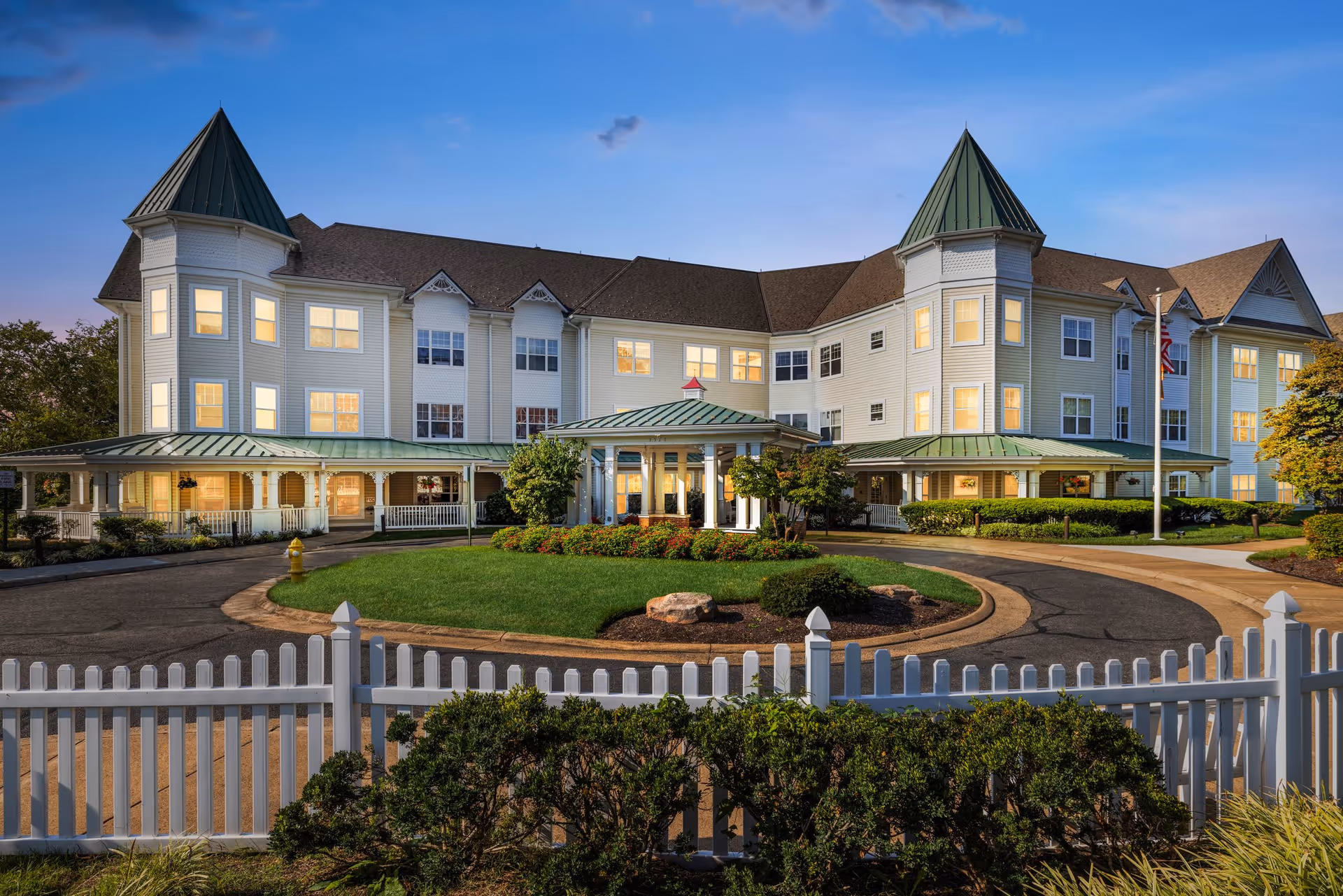 Exterior view of a large, multi-story senior living facility building with two prominent turret-like structures on each end, a circular driveway with a landscaped island in the center, and a white picket fence in the foreground during dusk with lights on inside the building.