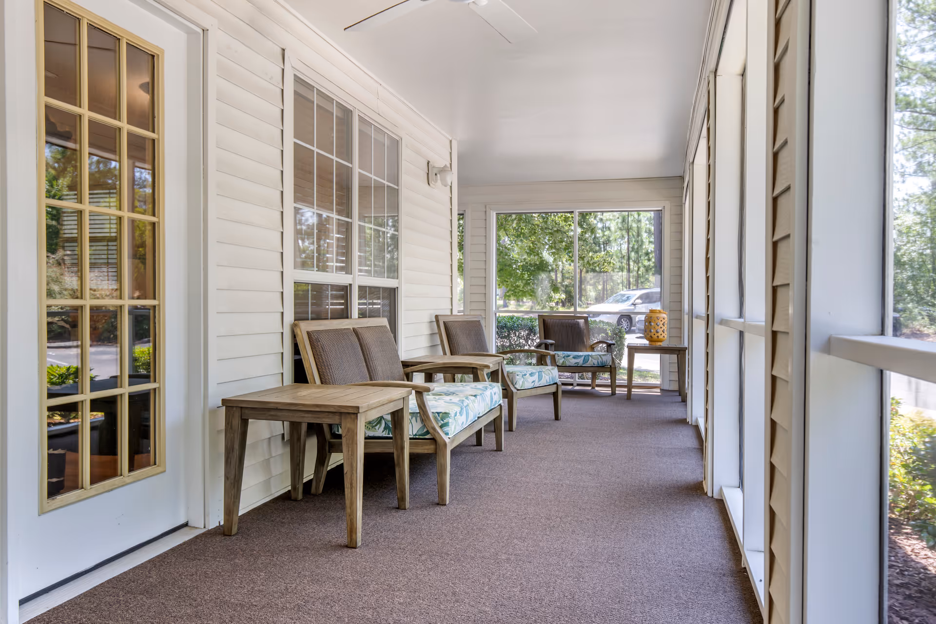 A screened-in porch area with three wooden chairs featuring floral cushions and two wooden side tables. The porch has beige siding, large windows, and a glass door with multiple panes. Outside, trees and a parked car are visible.