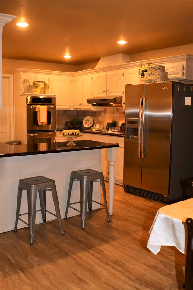 A warm, well-lit kitchen with white cabinetry, a stainless steel refrigerator, and a built-in oven. There is a black countertop island with two metal stools in front of it. The floor has a wood-like finish, and a table with a white tablecloth is partially visible on the right side.