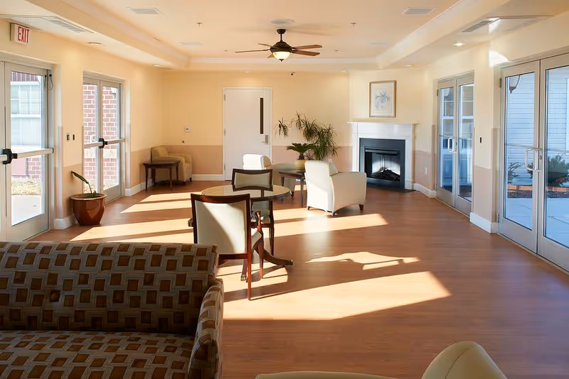 A bright and spacious living room area in an assisted living facility with wooden flooring, several chairs and tables, a fireplace, potted plants, and large glass doors letting in natural light.