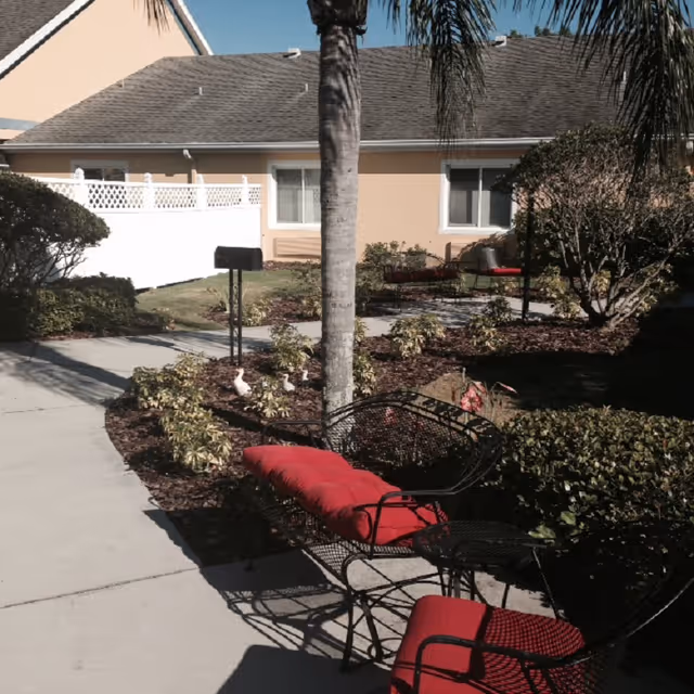 Outdoor patio area with black metal chairs and a bench featuring red cushions, surrounded by landscaped garden beds with small shrubs and a palm tree, adjacent to a beige building with white windows and a white fence.