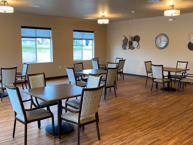 A spacious dining room with several square wooden tables and cushioned chairs arranged neatly. The room has wooden flooring, beige walls, two windows with black roller shades, ceiling lights, a wall clock, and decorative wall art.