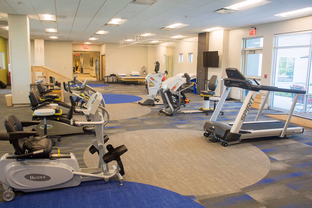 A spacious fitness room in a senior living facility featuring various exercise equipment including recumbent bikes, a treadmill, and strength training machines. The room has a modern design with patterned carpet, bright overhead lighting, and large windows allowing natural light. There are also therapy tables and a mounted TV on the wall.