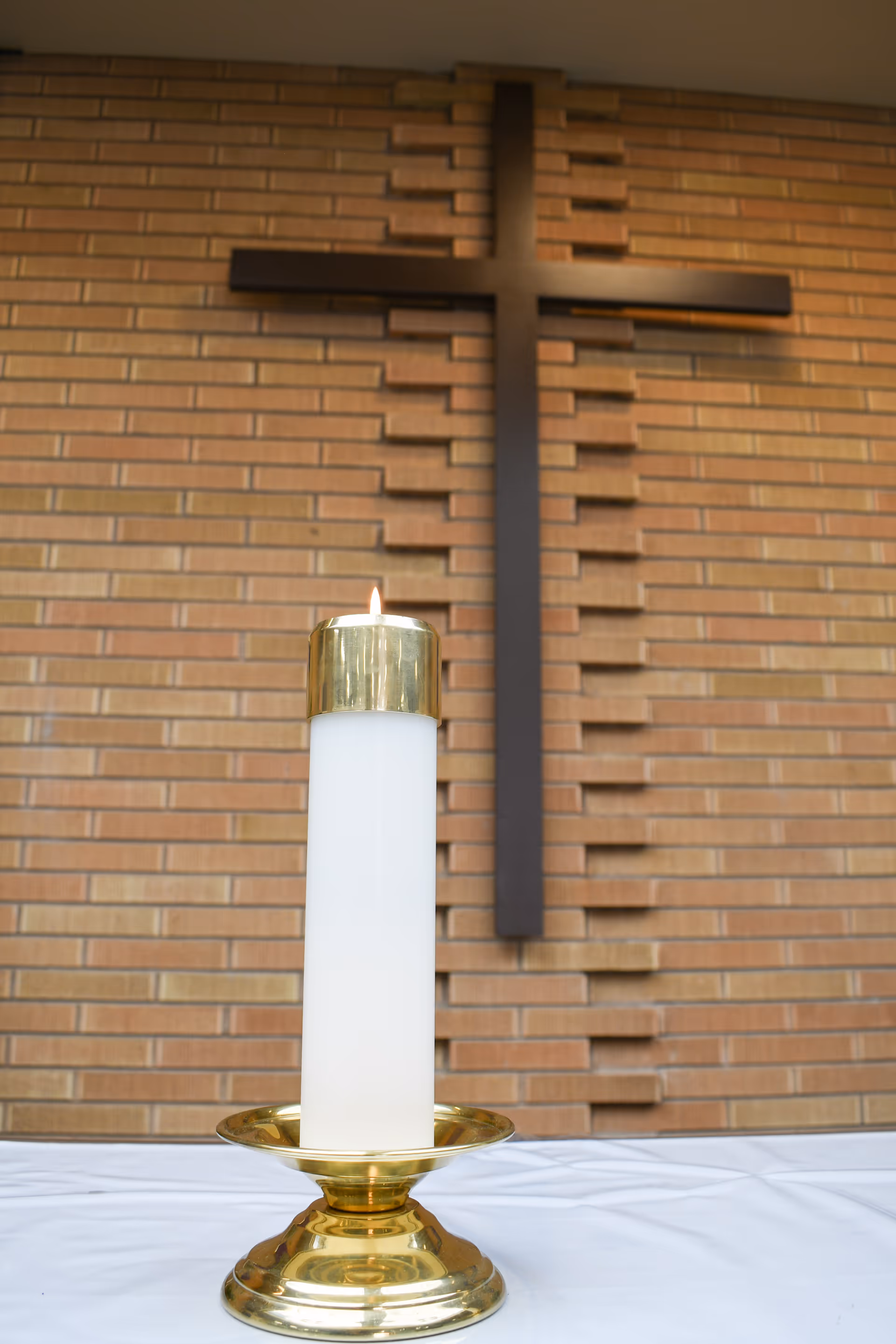 A lit white candle with a gold holder placed on a white cloth-covered surface in front of a brick wall with a large dark wooden cross mounted on it.