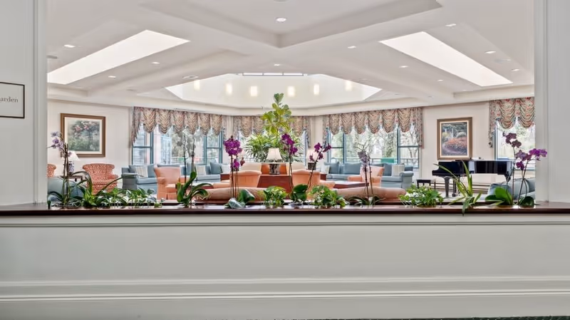A bright and spacious living room area with large windows allowing natural light to fill the space. The room features comfortable seating including sofas and armchairs arranged around a central table with a lamp and decorative plants. There are floral curtains on the windows, framed artwork on the walls, and a grand piano in the corner. The ceiling has recessed lighting and a skylight, and there is a planter with green plants and purple orchids in the foreground.