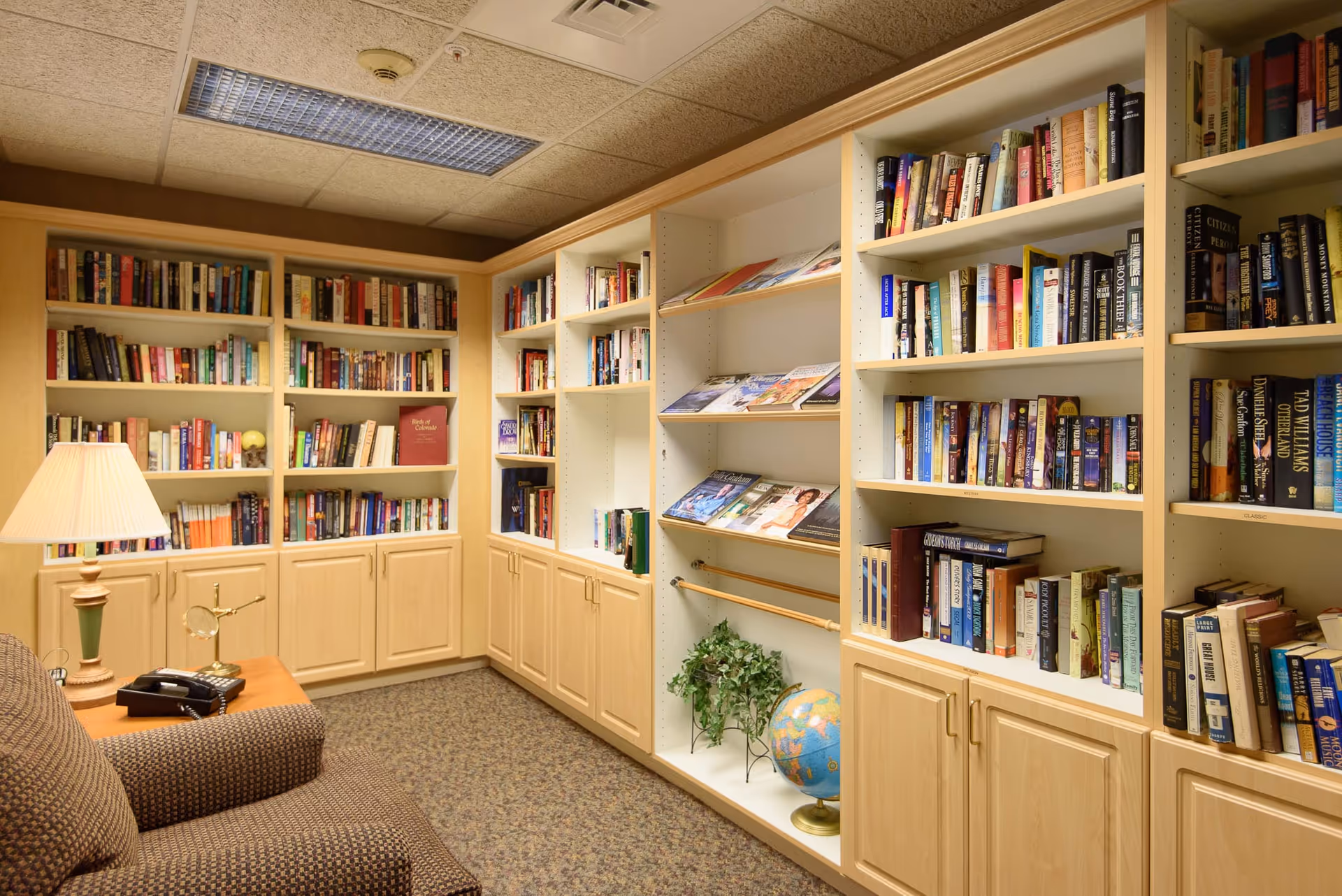 A cozy library room with built-in wooden bookshelves filled with books and magazines. There is a comfortable upholstered armchair next to a wooden side table with a lamp and a telephone. The carpeted floor and ceiling tiles give the room a warm, inviting atmosphere.