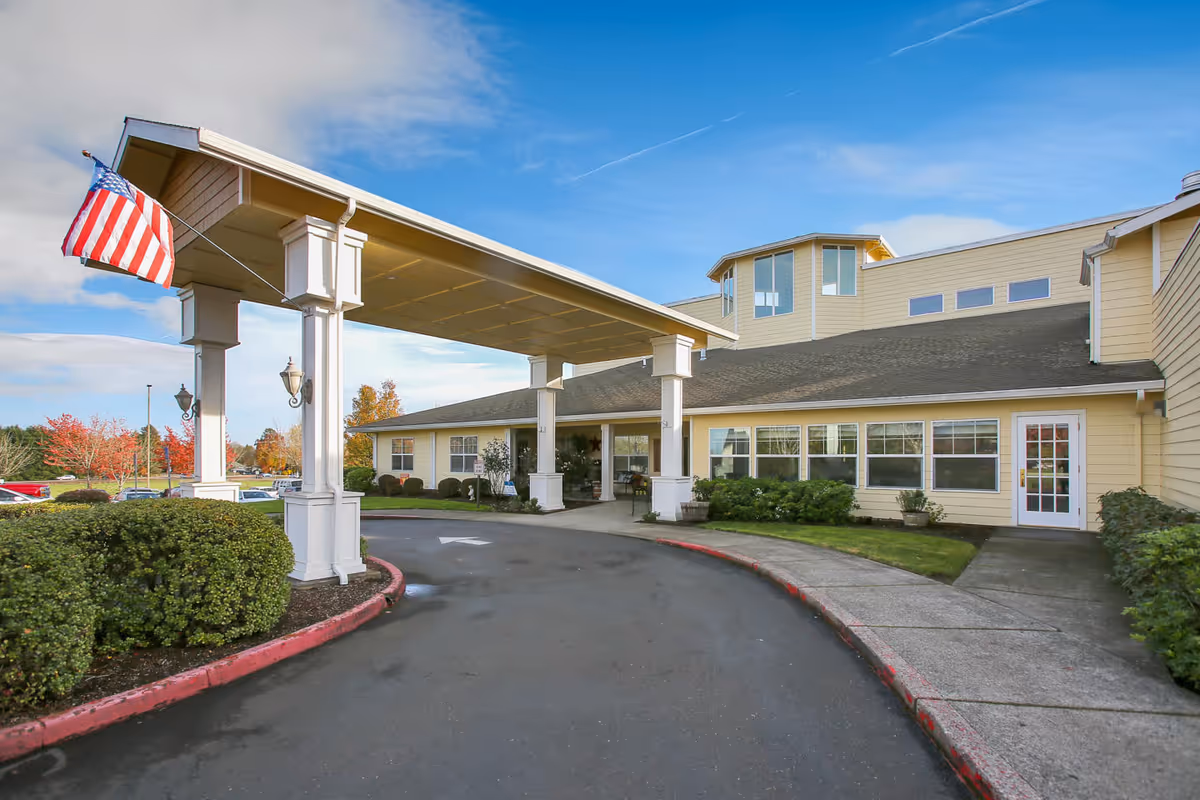 Front entrance of a yellow senior living facility with a covered porte-cochere, American flag, and landscaped driveway.