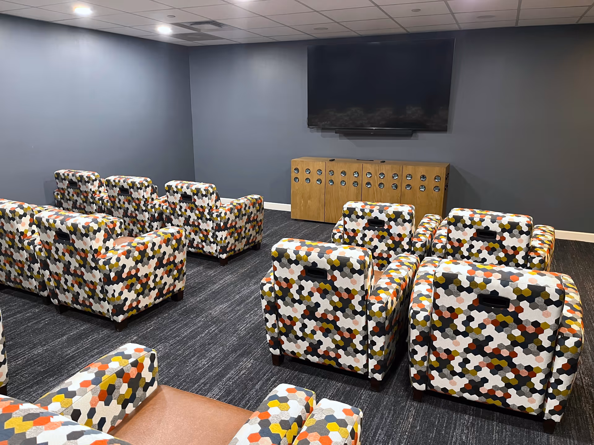 A small theater or media room with multiple armchairs arranged in rows facing a large flat-screen TV mounted on a gray wall. The armchairs have a colorful hexagonal pattern in shades of white, black, orange, yellow, and gray. Below the TV is a wooden cabinet with circular cutouts.