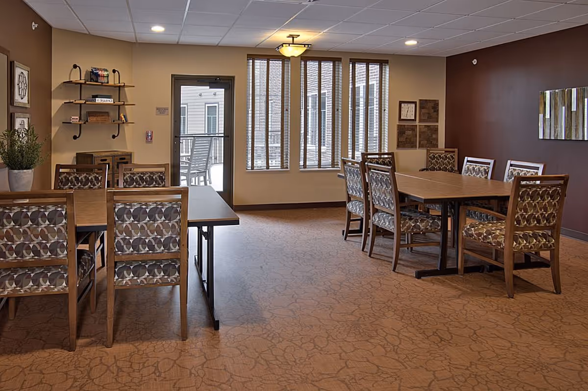 A dining room with two wooden tables surrounded by patterned cushioned chairs. The room has beige walls with a darker accent wall, a carpeted floor, a door leading outside, and windows with vertical blinds letting in natural light. There are decorative shelves and framed artwork on the walls.
