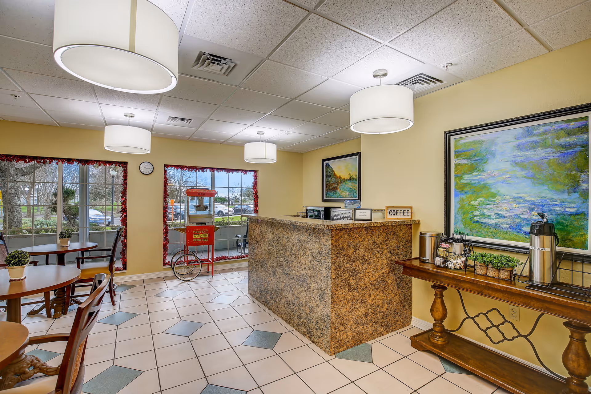 A cozy common area with a granite countertop serving station labeled 'COFFEE', a popcorn machine near large windows decorated with red garlands, round wooden tables with chairs, and a wooden side table holding coffee supplies and small potted plants. The walls are painted yellow and adorned with colorful paintings. The floor has a tiled pattern with white and green tiles, and the ceiling has white hanging light fixtures.