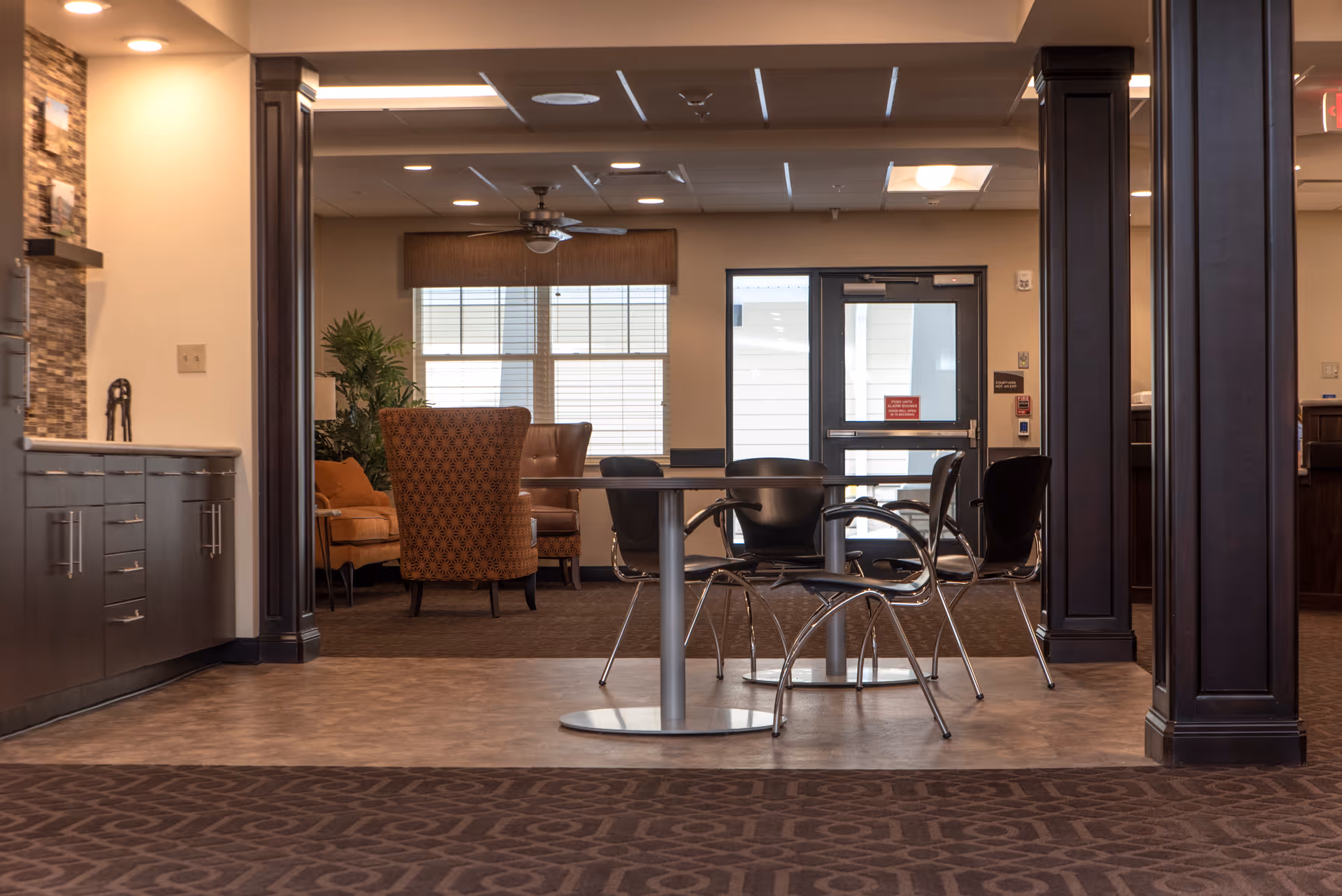 Interior view of a senior living facility lounge area with a round table and four black chairs in the foreground. In the background, there are two upholstered armchairs and a sofa near a window with blinds. The room features dark wood columns, a ceiling fan, and a door leading outside.