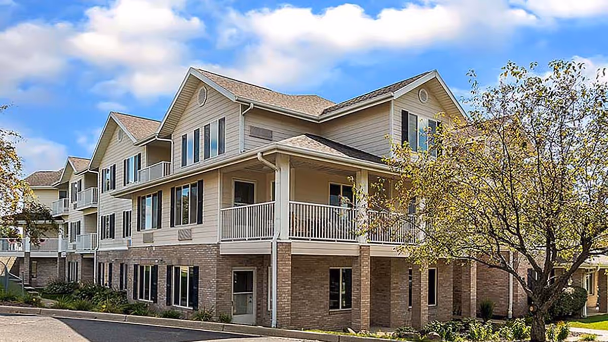 Exterior view of a multi-story residential building with beige siding and brick accents, featuring balconies and surrounded by trees under a partly cloudy sky.