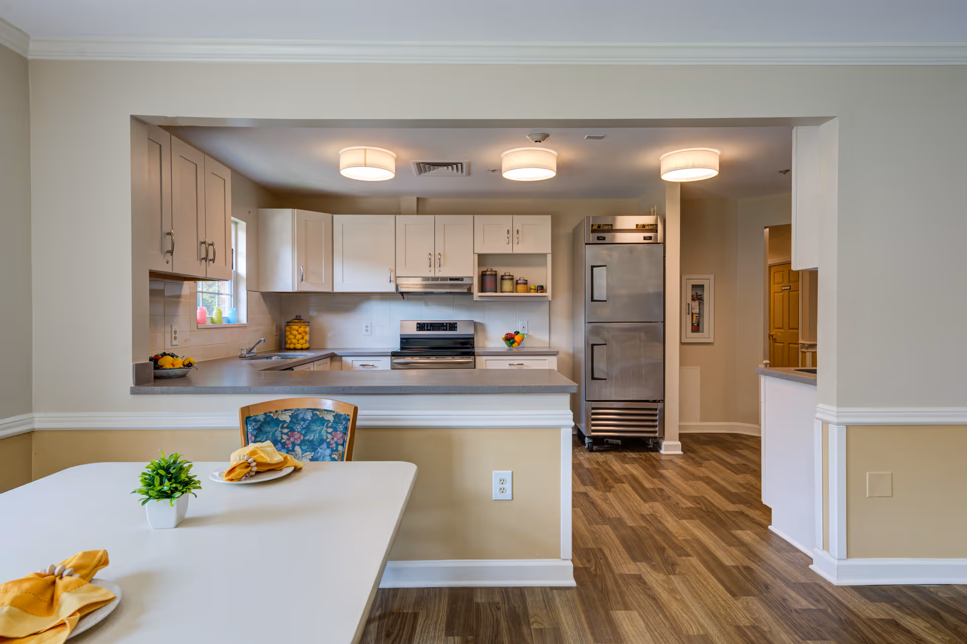 A clean and modern kitchen area with white cabinets, a stainless steel refrigerator, stove, and countertop. In the foreground, there is a dining table with a small green plant and yellow napkins on plates. The floor has wood-patterned flooring, and the walls are painted in neutral tones with white trim.