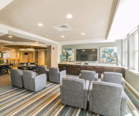 A spacious common area in a senior living facility with several gray upholstered armchairs arranged facing a large flat-screen TV mounted on a wall with two abstract paintings on either side. The room has large windows letting in natural light, a striped carpet, and additional seating and tables in the background.