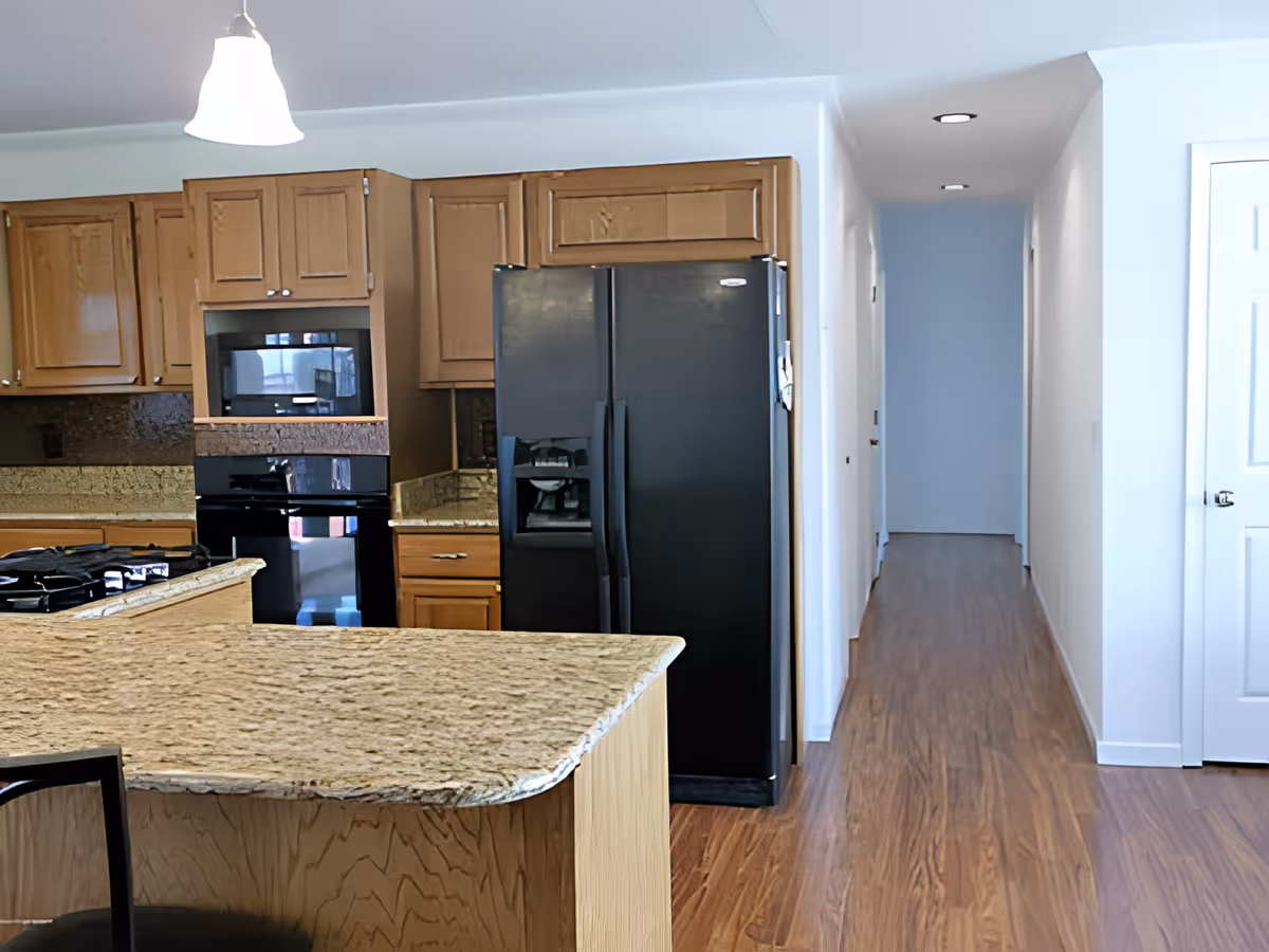 Open kitchen with a granite island, wooden cabinets, black refrigerator and built-in appliances, and a hallway to the right.