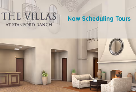 Interior view of a senior living facility lounge area with a fireplace, two armchairs, a coffee table, potted plants, and a reception desk in the background. The space has high ceilings and modern light fixtures.