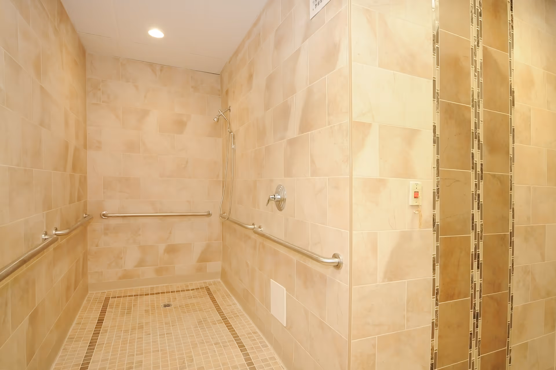 A spacious walk-in shower with beige tiled walls and floor, equipped with stainless steel grab bars on three sides and a handheld showerhead. The shower area is well-lit with a recessed ceiling light.