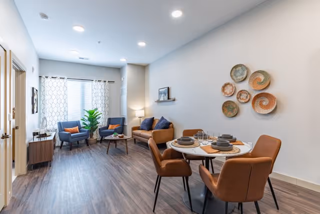A modern living and dining area with wood flooring, featuring a round dining table set with four brown chairs and place settings. The living area has a brown sofa, two blue armchairs with orange cushions, a small coffee table, a floor lamp, and a large window with patterned curtains. Decorative woven baskets are mounted on the wall above the dining table.