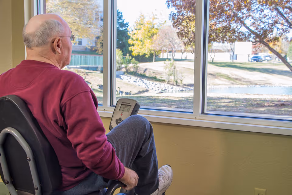 An elderly man wearing a maroon sweatshirt and gray pants is seated on an exercise bike indoors, looking out through a large window at a scenic outdoor area with trees and a small pond.