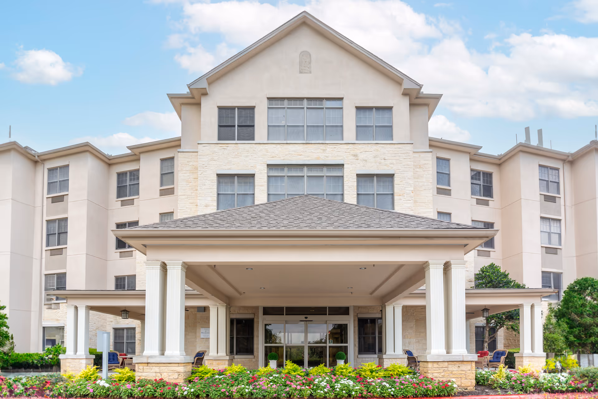 Front entrance of a multi-story beige senior living building with a covered portico and landscaped flower beds.