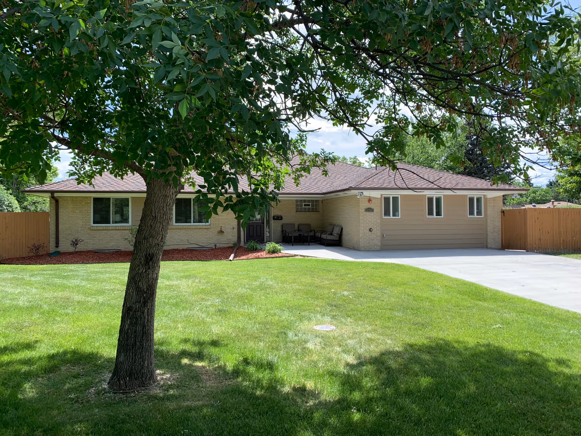 Front exterior view of a single-story residential building with a well-maintained green lawn and a large tree in the foreground. The house has a light-colored brick facade, a brown shingled roof, and a driveway leading to a garage on the right side. There is a small covered porch area with outdoor seating near the front door.