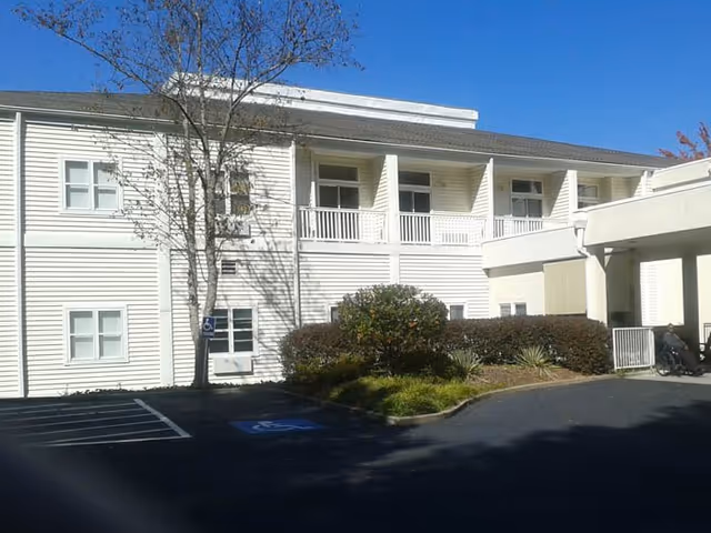 White two-story senior living building with parking, landscaping, and a wheelchair-accessible entrance.
