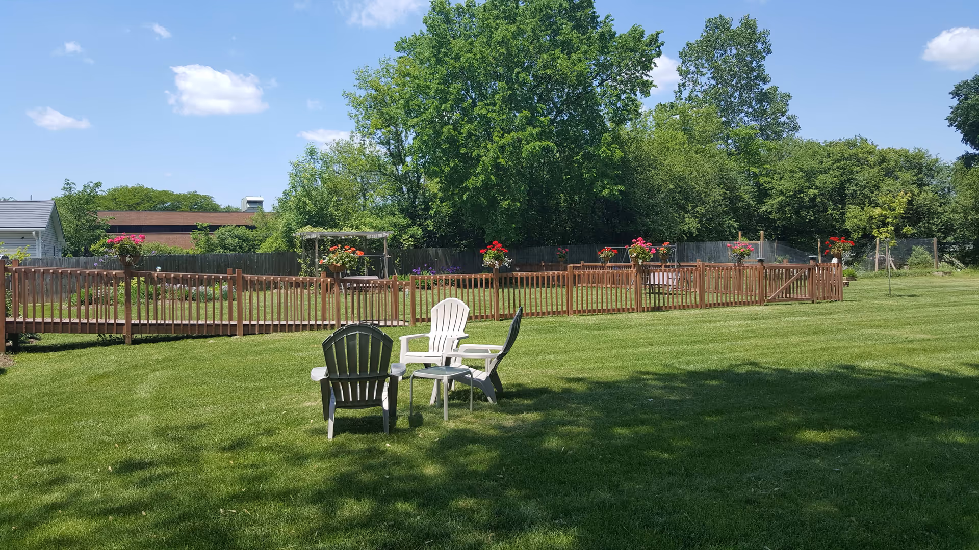 A sunny outdoor area with green grass, three plastic chairs arranged around a small table, a wooden fence with flower pots on it, and trees in the background under a blue sky with a few clouds.