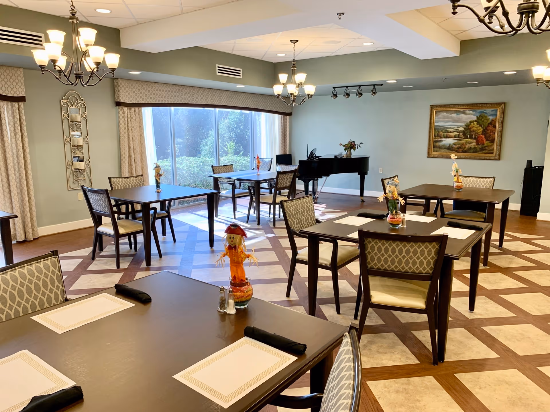 A bright dining room with several dark wood tables and cushioned chairs arranged neatly. Each table has placemats, black napkins, and small decorative centerpieces. Large windows with curtains allow natural light to fill the room. A black grand piano is positioned against one wall, and a framed landscape painting hangs nearby. The floor has a patterned tile design, and chandeliers hang from the ceiling.