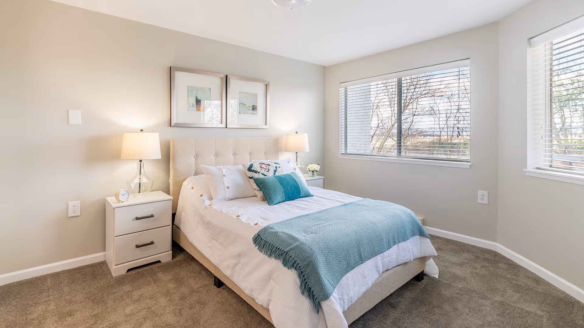 A bright and cozy bedroom with a neatly made bed featuring white bedding and teal accent pillows and throw. There are two bedside tables with matching lamps on either side of the bed. Two framed pictures hang above the headboard. Large windows with blinds allow natural light to fill the room, and the floor is carpeted in a neutral tone.