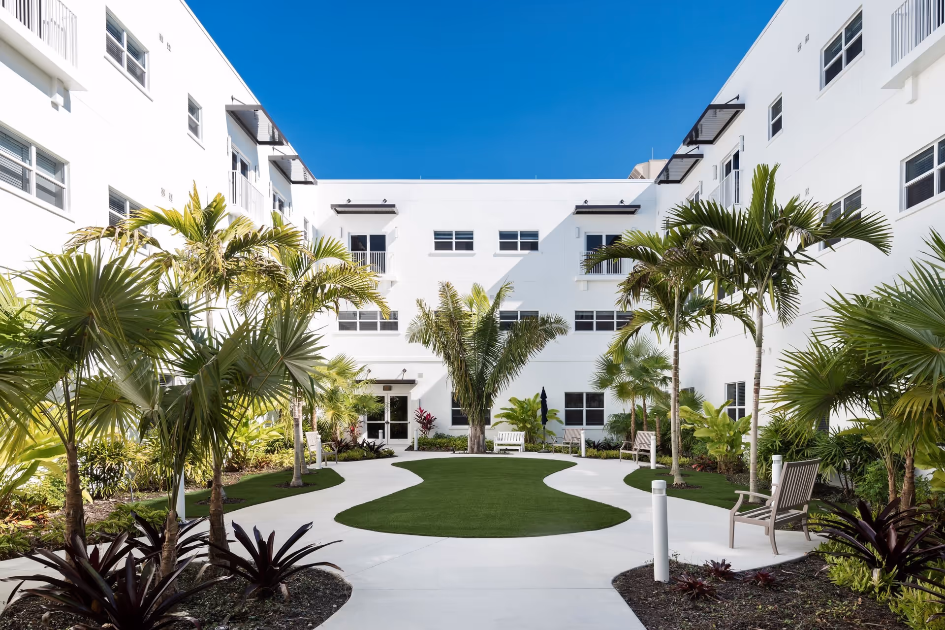 Outdoor courtyard area at Amavida at Lakes Park featuring a curved concrete walkway surrounding a green lawn, with palm trees and various plants along the sides. White benches are placed along the walkway, and the courtyard is enclosed by a white multi-story building under a clear blue sky.
