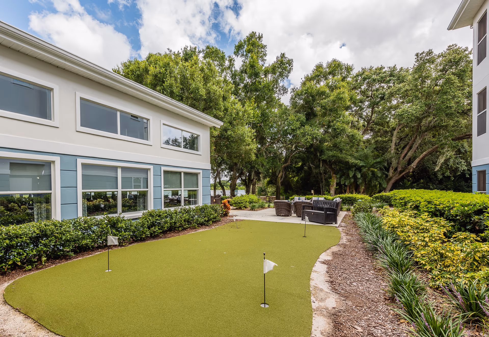 Outdoor putting green area surrounded by bushes and trees, with a seating area featuring wicker chairs and a bench near a two-story building with large windows under a partly cloudy sky.