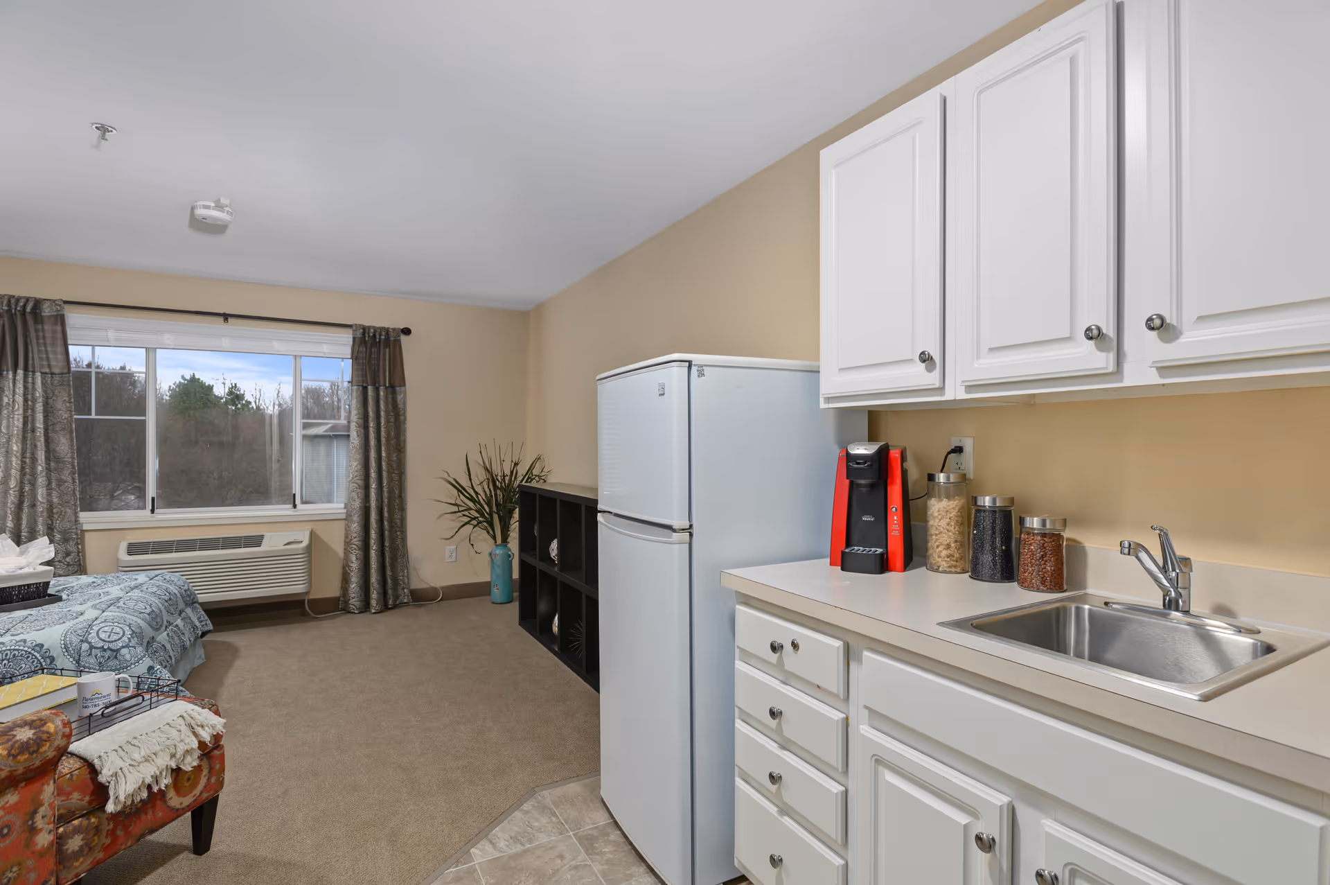 Interior view of a senior living facility room showing a small kitchenette with white cabinets, a sink, a red coffee maker, and jars of pasta and beans on the counter. Next to the kitchenette is a white refrigerator. The room has beige walls and carpeted floor, with a window covered by patterned curtains. A bed with a blue patterned bedspread and a colorful upholstered bench are partially visible.