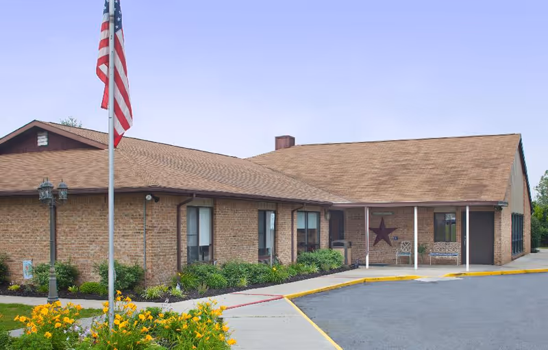 Exterior view of a single-story brick building with a brown shingled roof, an American flag on a flagpole, a flower bed with yellow flowers, and a sidewalk leading to the entrance with a large star decoration on the wall and benches nearby.
