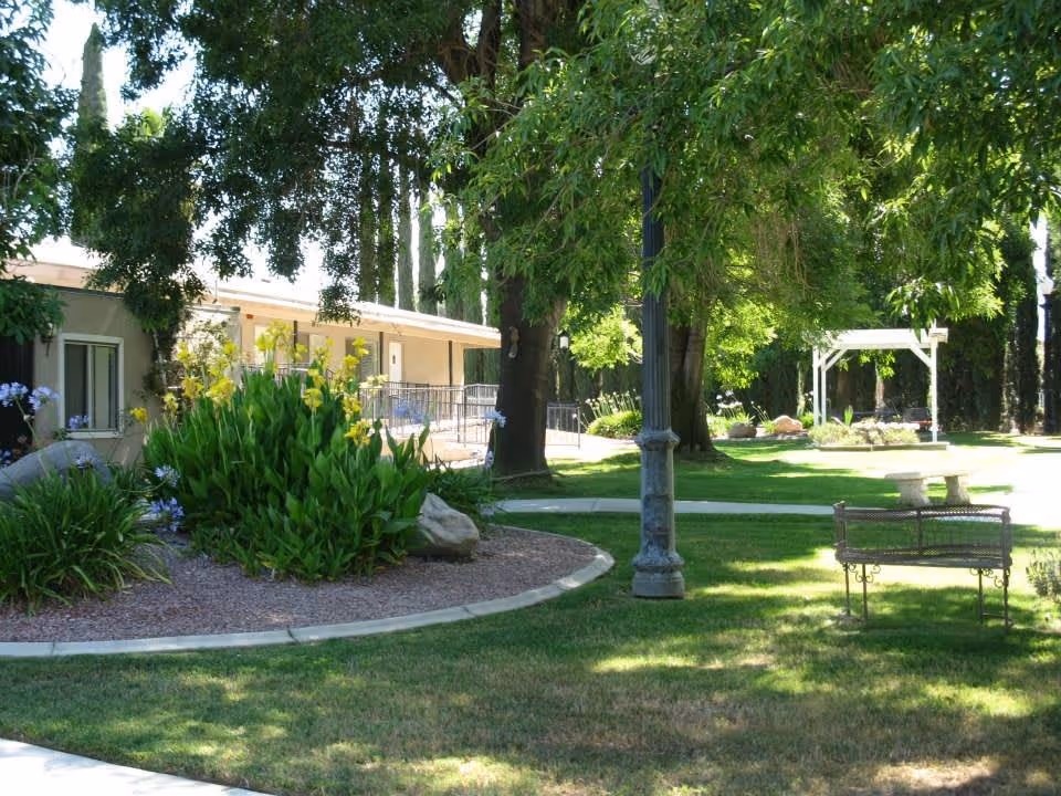 A peaceful outdoor garden area with green grass, trees providing shade, flowering plants, a stone bench, a metal bench, and a pathway. A building with a covered porch is visible in the background.