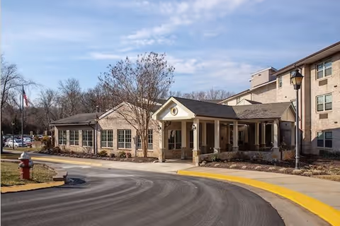 Exterior view of a multi-story brick building with a covered entrance and a curved driveway. There are leafless trees, a fire hydrant, and a street lamp visible under a partly cloudy sky.