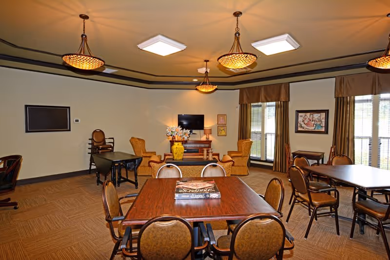 A well-lit common room with multiple tables and chairs arranged for group seating. The room features a television on a wooden stand, two armchairs, a yellow vase with flowers, and large windows with brown curtains allowing natural light to enter. The ceiling has decorative light fixtures and recessed lighting.