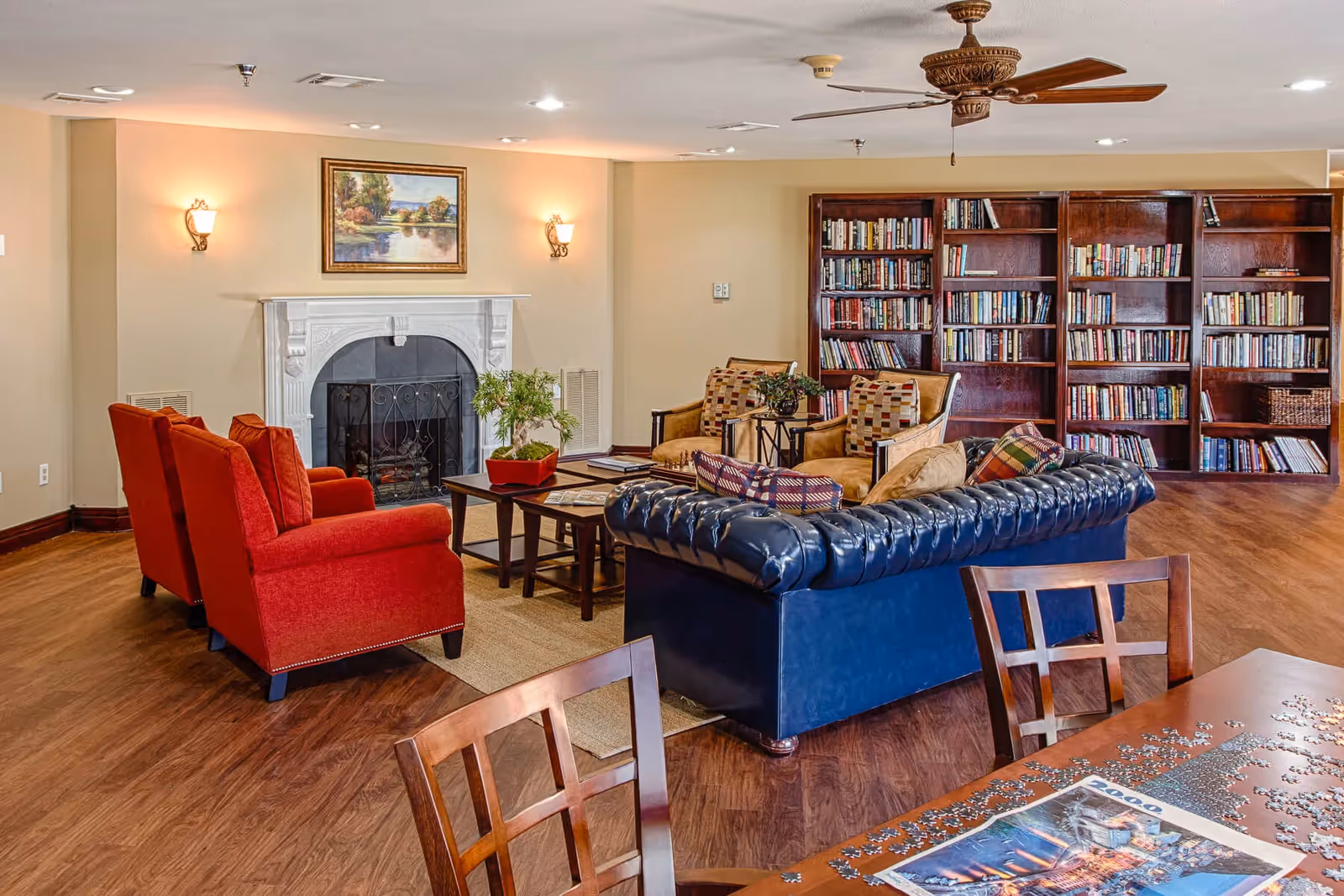 Cozy common lounge with a fireplace, large bookshelf, a blue leather sofa and red armchairs arranged around a coffee table.