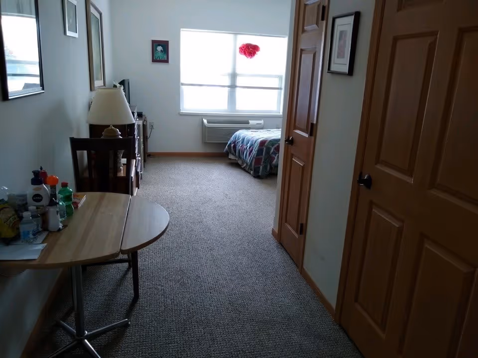 View down a carpeted hallway in a senior living facility room with wooden doors on the right, a small wooden table with various personal items and a chair on the left, and a bed with a colorful quilt near a window at the far end. A lamp and framed pictures are visible on the left wall.