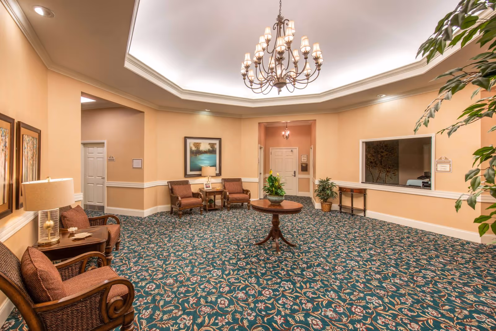 A spacious, well-lit common area with beige walls and a patterned green carpet. The room features a round wooden table with a plant in the center, several cushioned chairs with wooden frames, framed artwork on the walls, and a chandelier hanging from a recessed ceiling. There are doorways and a windowed reception area visible in the background.